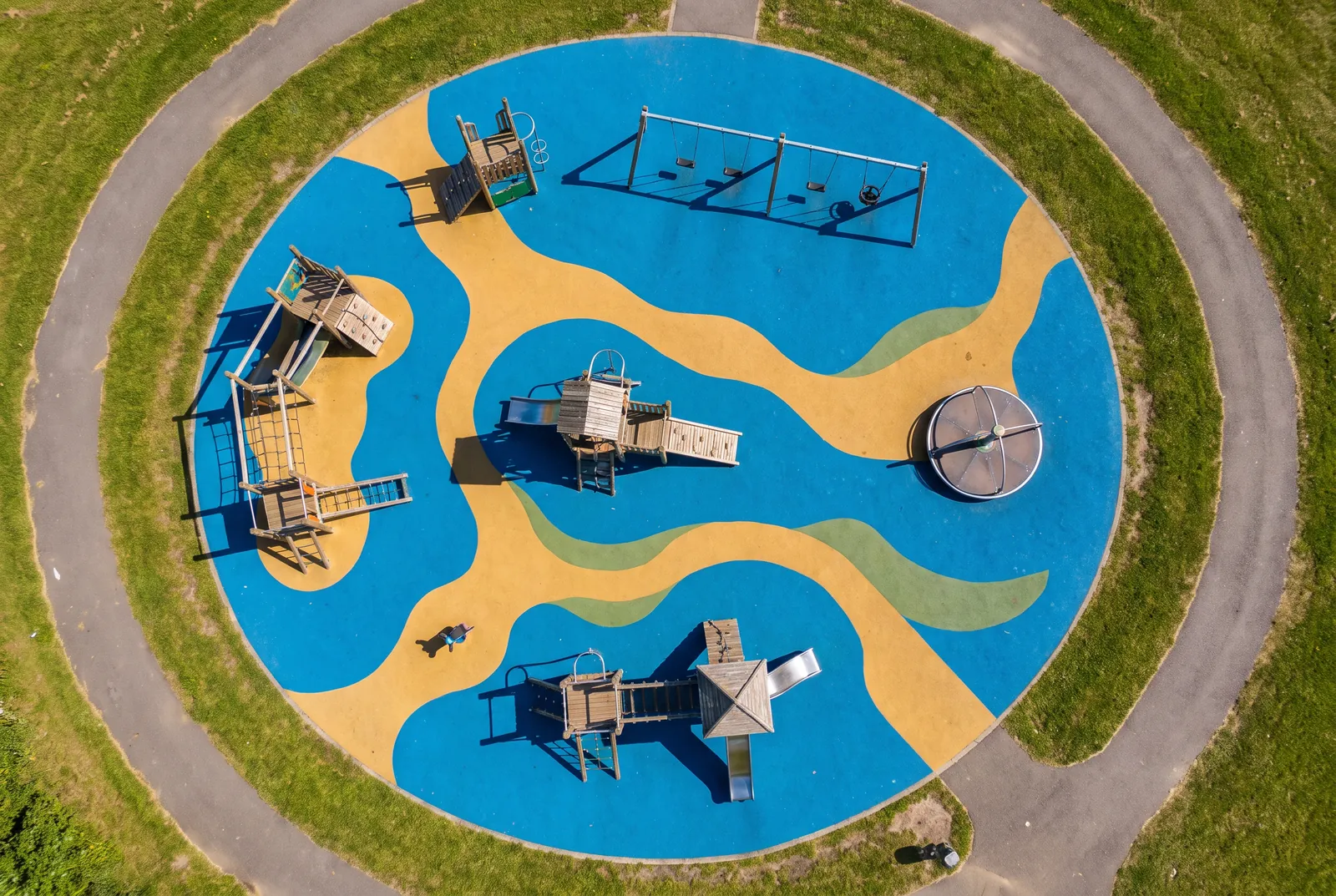 An aerial view of a playground surfaced in blue and gold poured rubber shapes around play structures, swings and a spinner