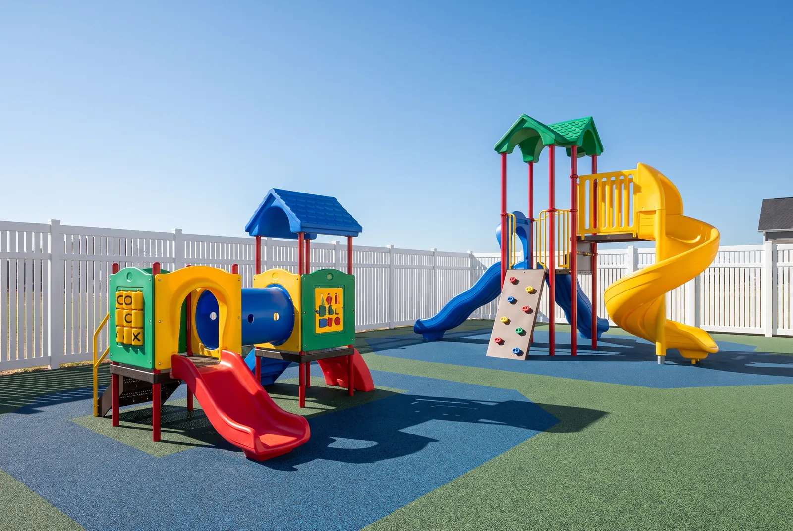 Two play structures inside a white fence at a child care center: a toddler structure with a short slide and crawl-through, and a preschool structure with a spiral slide and climbing wall