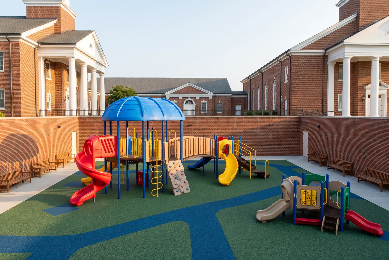A church courtyard playground between brick buildings with white columns: a play structure with a blue roof, red and yellow slides and a small toddler structure beside it