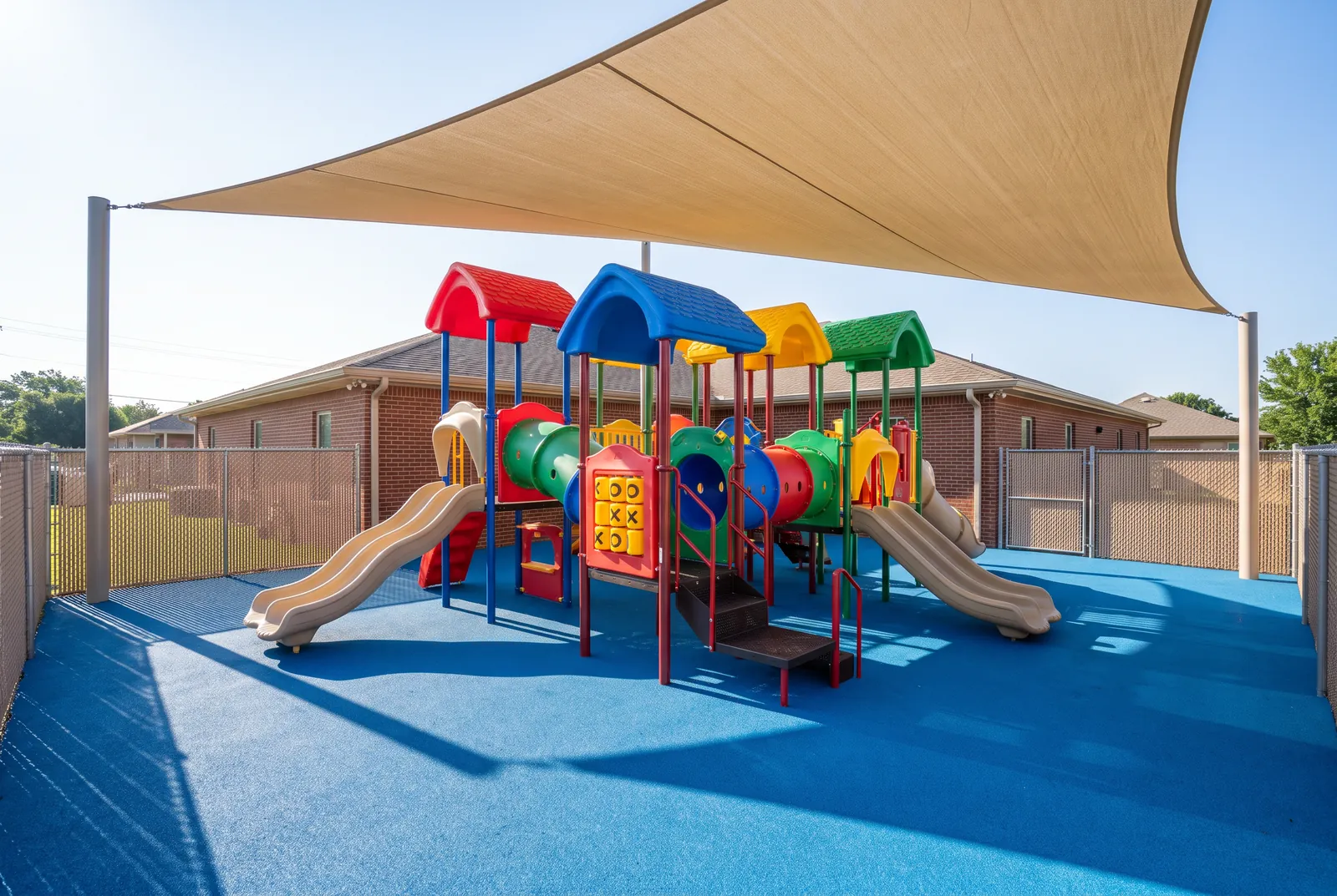A daycare playground behind a brick child care building: a preschool play structure with red, blue, yellow and green roofs, slides and a crawl tunnel on blue rubber under a shade sail