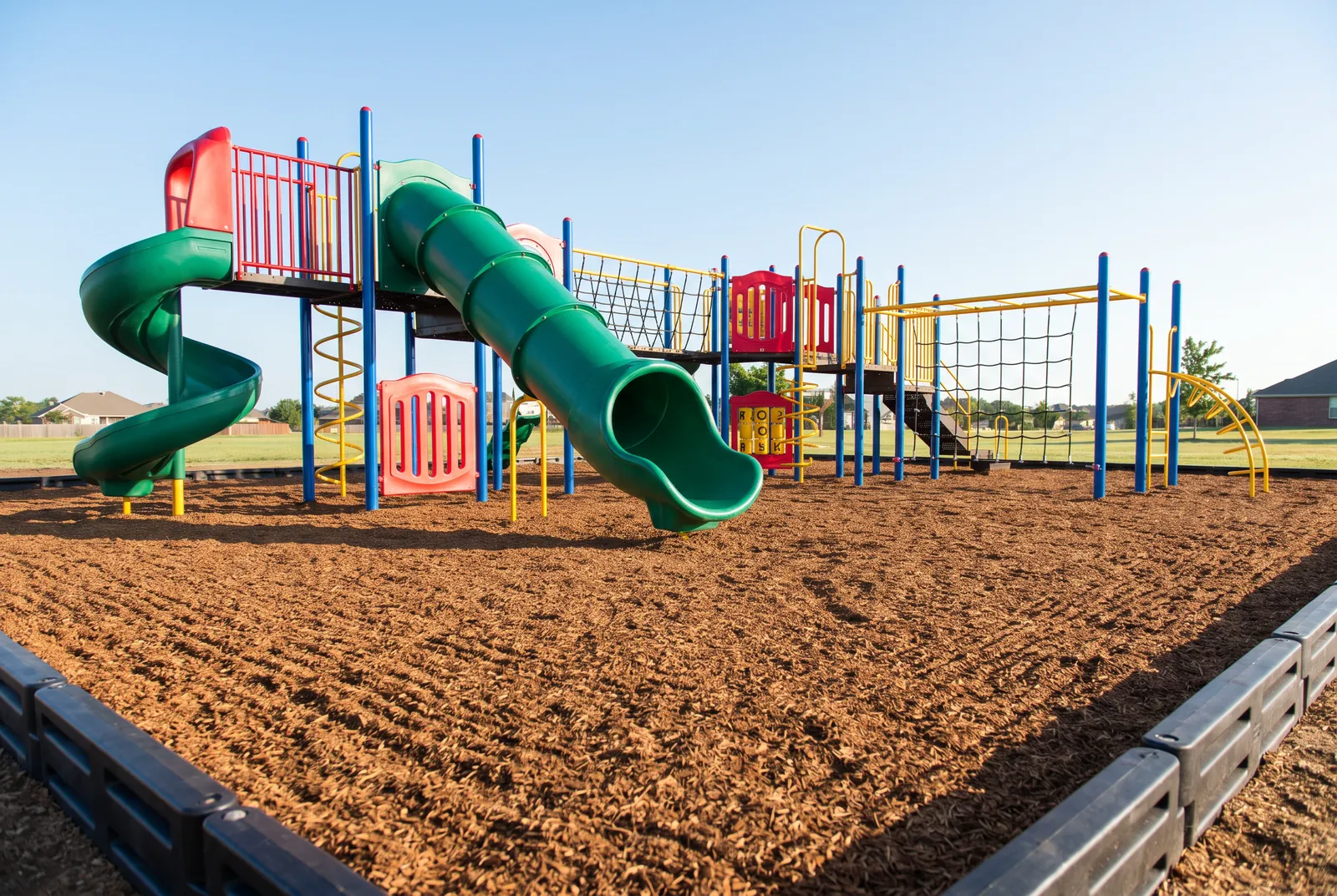 A thick, freshly raked bed of engineered wood fiber inside a black border under a colorful play structure with green tube and spiral slides