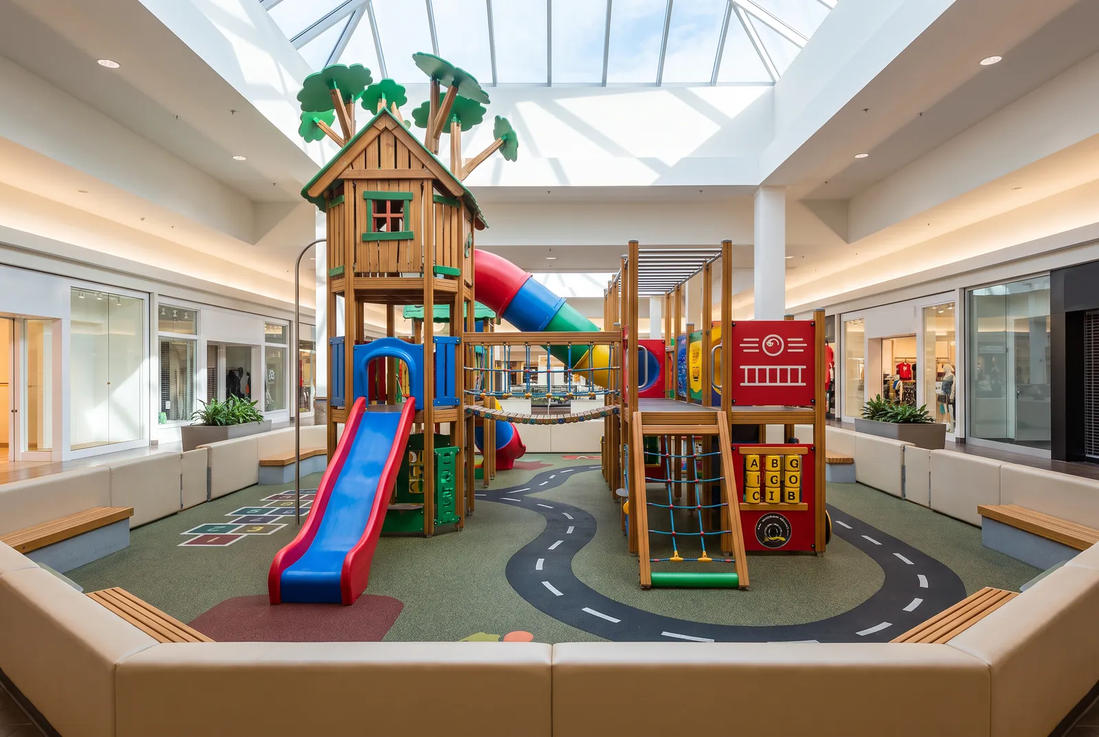 An indoor mall playground built like a treehouse with a fire-engine front, slides, nets and play panels on cushioned flooring with a painted road