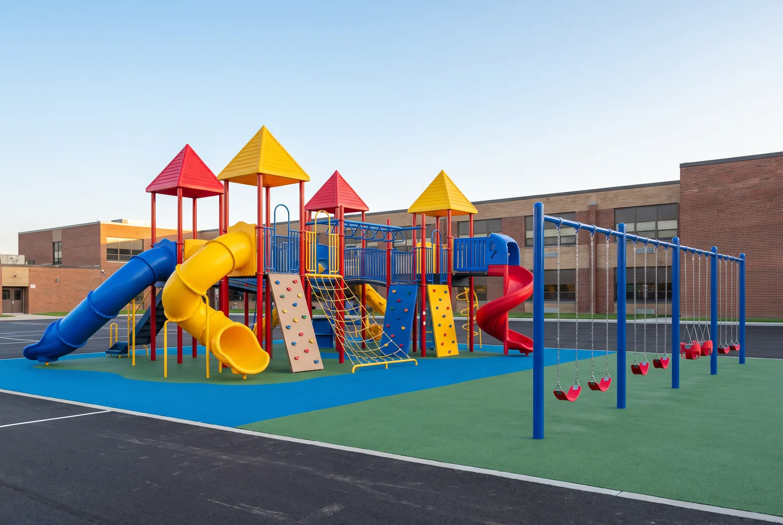 A school playground with red and yellow roofs, tube and spiral slides, climbing walls and a row of swings on blue and green poured rubber