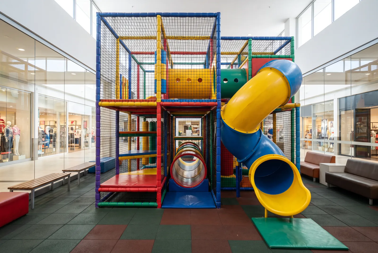 A two-level indoor soft contained play structure with netting, a yellow and blue spiral slide and crawl tunnels in a shopping center