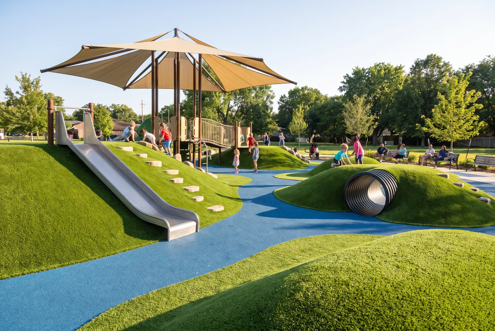 Green synthetic turf shaped into play mounds with a tunnel and an embankment slide, blue rubber paths and a shade sail